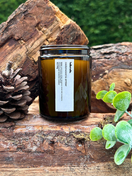 Cedarwood & Amber 150g scented candle by Hellomelts displayed on a natural wood log with pinecones and greenery, styled as a warm autumn lifestyle scene