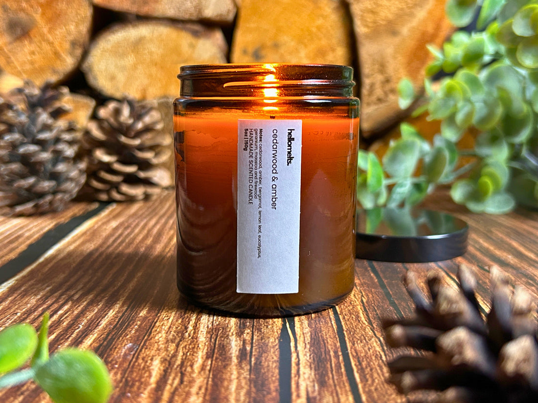 Lit Cedarwood & Amber candle from Hellomelts Autumn Collection 2025, in a amber glass jar on a rustic wooden surface with pinecones and greenery next to wooden logs