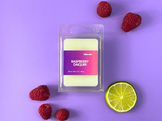 Hellomelts Raspberry Daiquiri wax melt from the 2025 Summer Collection, displayed in 85g clear clamshell packaging with white wax, surrounded by raspberries and a lime slice on a light purple background 
