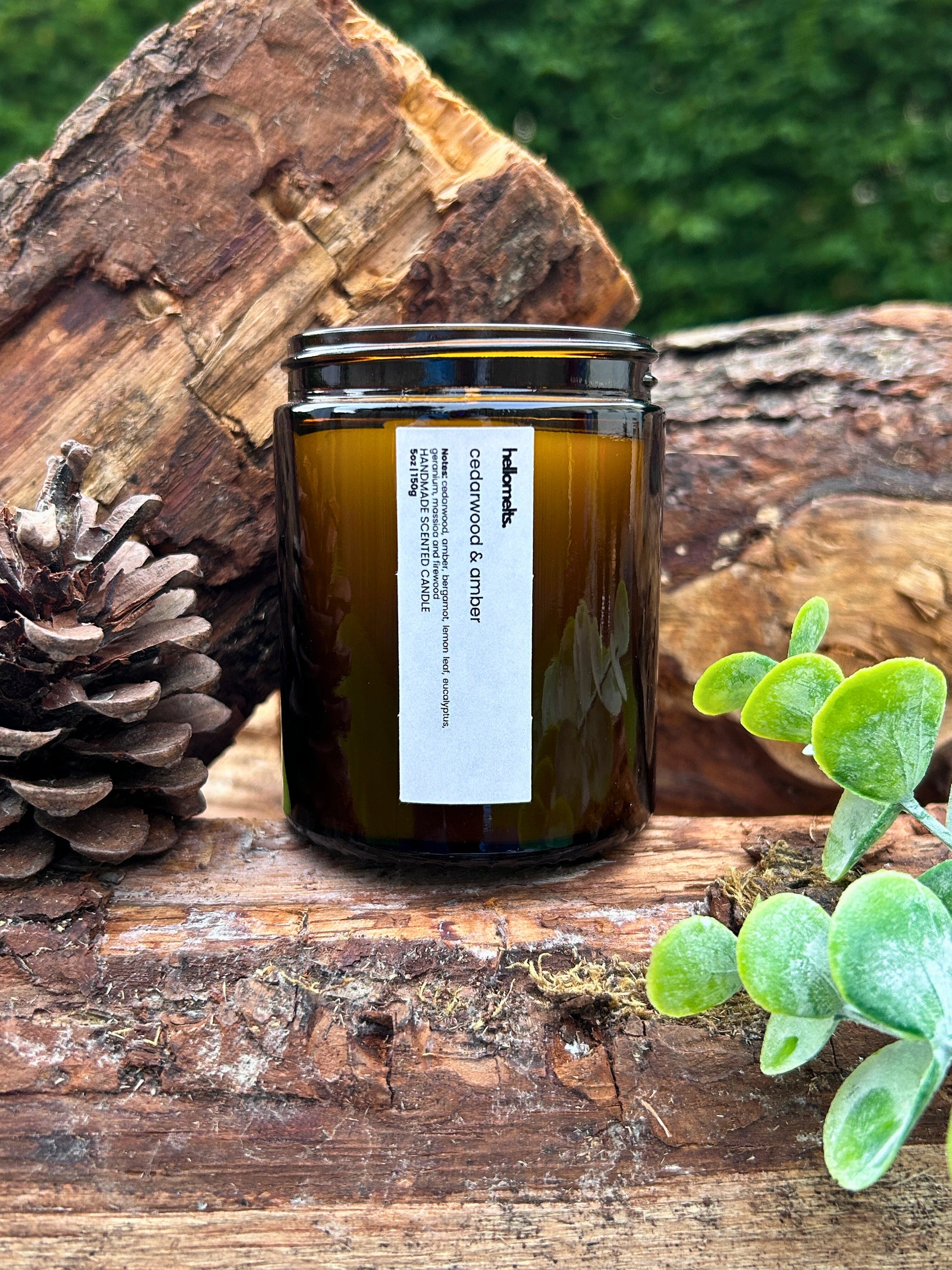 Cedarwood & Amber 150g scented candle by Hellomelts displayed on a natural wood log with pinecones and greenery, styled as a warm autumn lifestyle scene