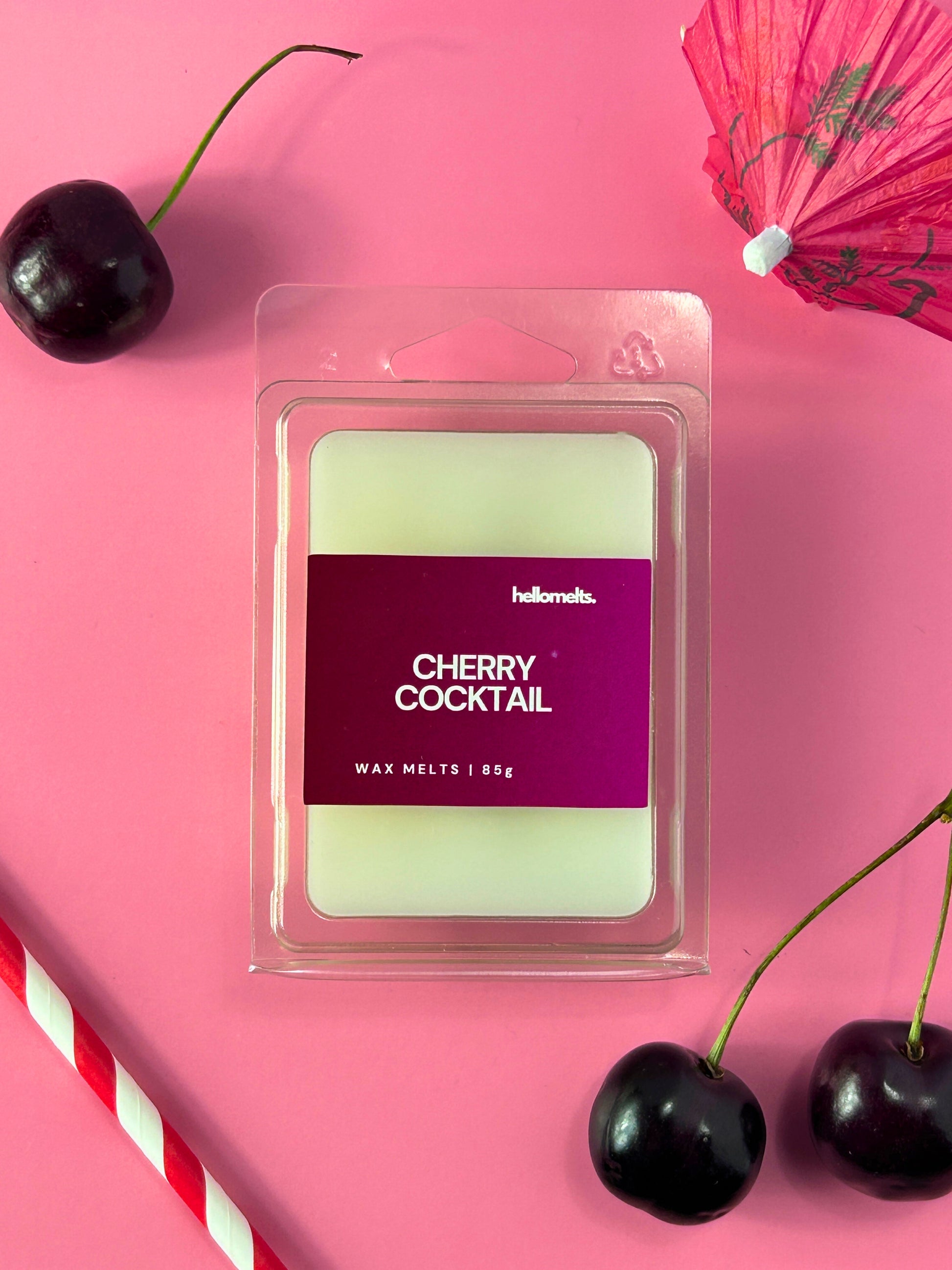 Cherry Cocktail wax melts by Hellomelts displayed in clear clamshell packaging on a pink background with fresh cherries, a red-and-white striped straw, and a cocktail umbrella, part of the Summer Collection