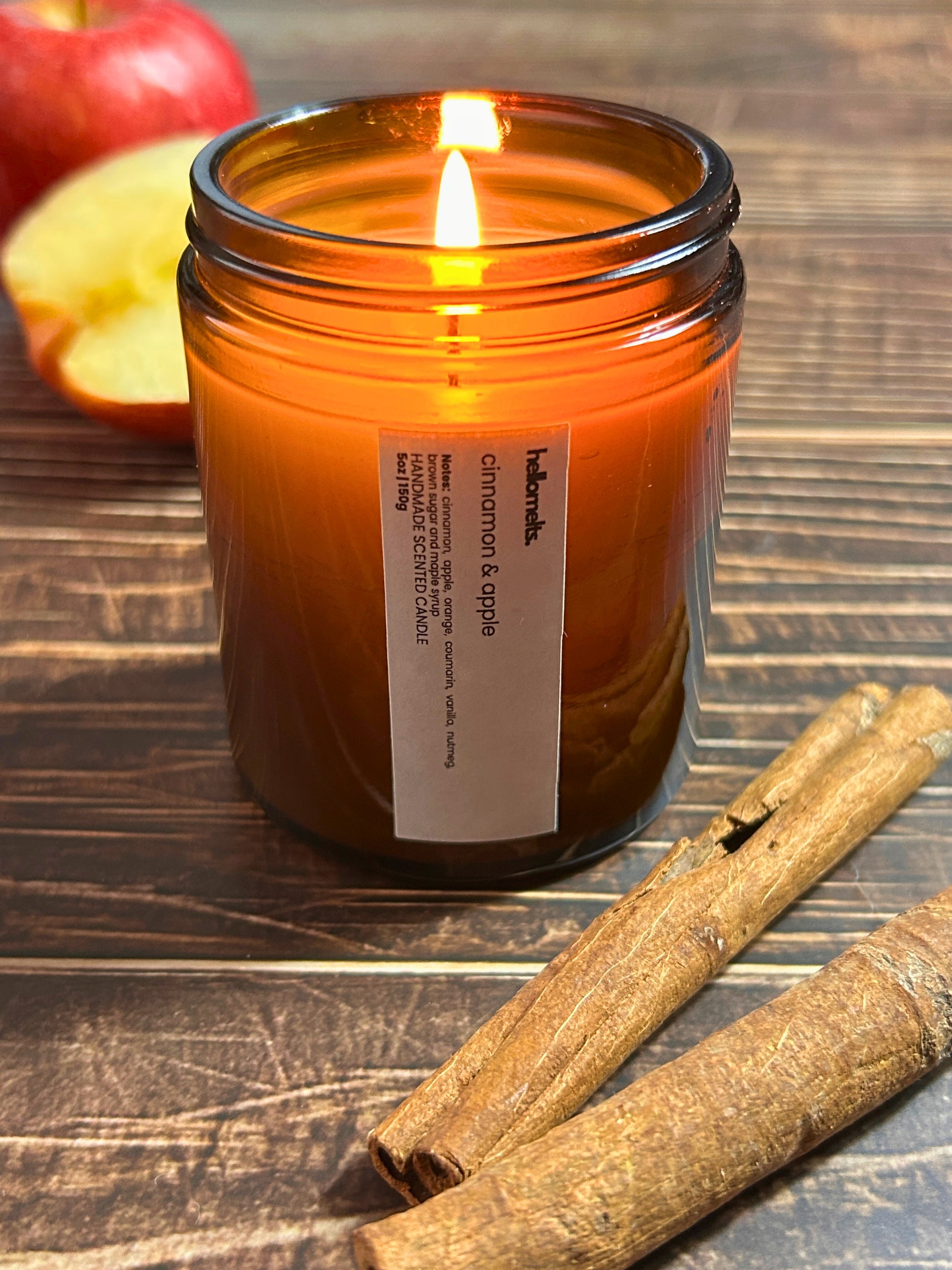 Cinnamon & Apple scented candle by Hellomelts, lit and glowing in a amber glass jar surrounded by fresh apple and cinnamon sticks, styled as a cosy autumn lifestyle scene
