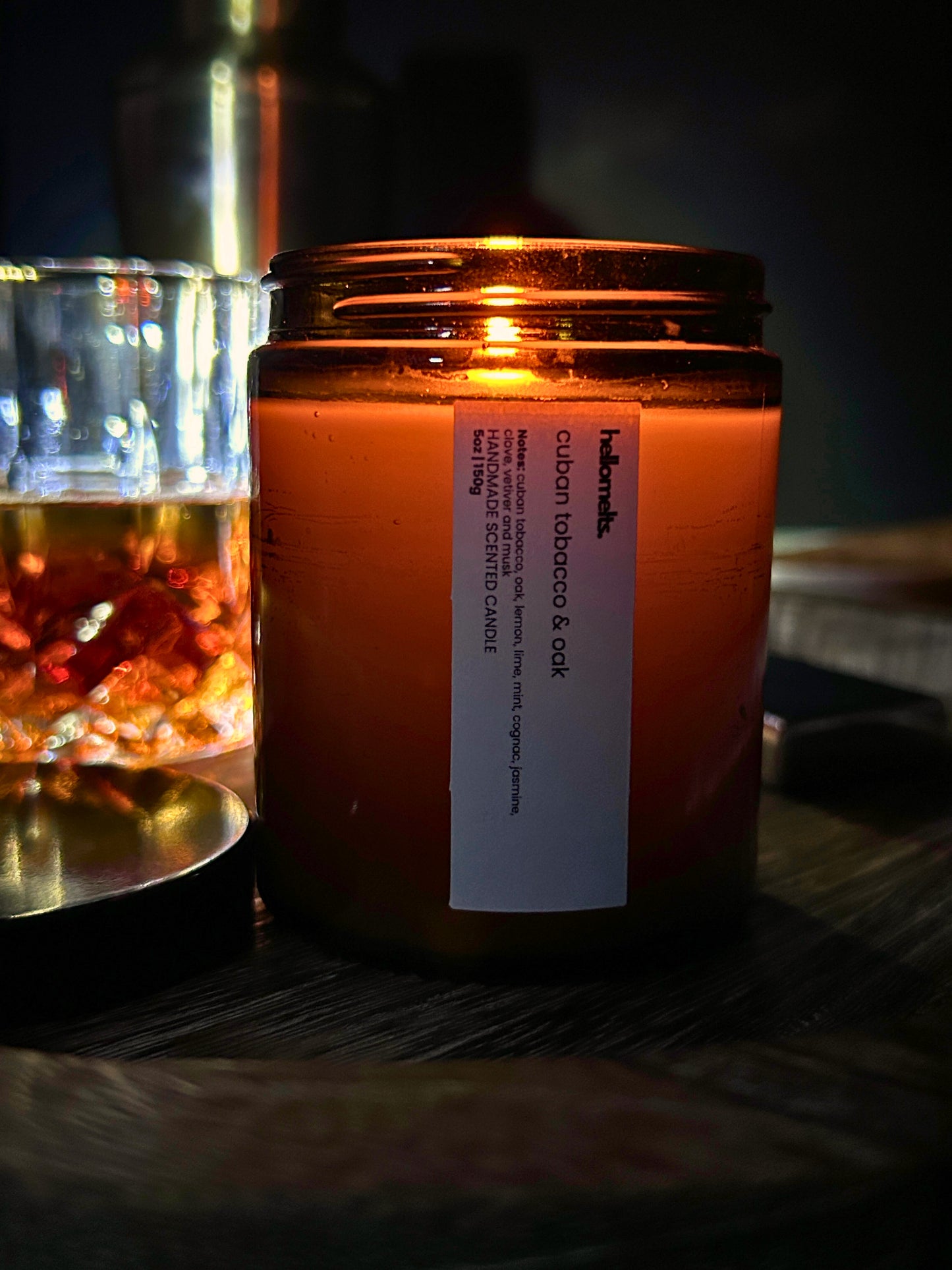 Cuban Tobacco & Oak 150g scented candle by Hellomelts, shown in an autumn lifestyle setting with warm amber glow, glass tumbler and wooden surface with candle lit and burning