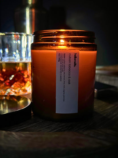 Cuban Tobacco & Oak 150g scented candle by Hellomelts, shown in an autumn lifestyle setting with warm amber glow, glass tumbler and wooden surface with candle lit and burning