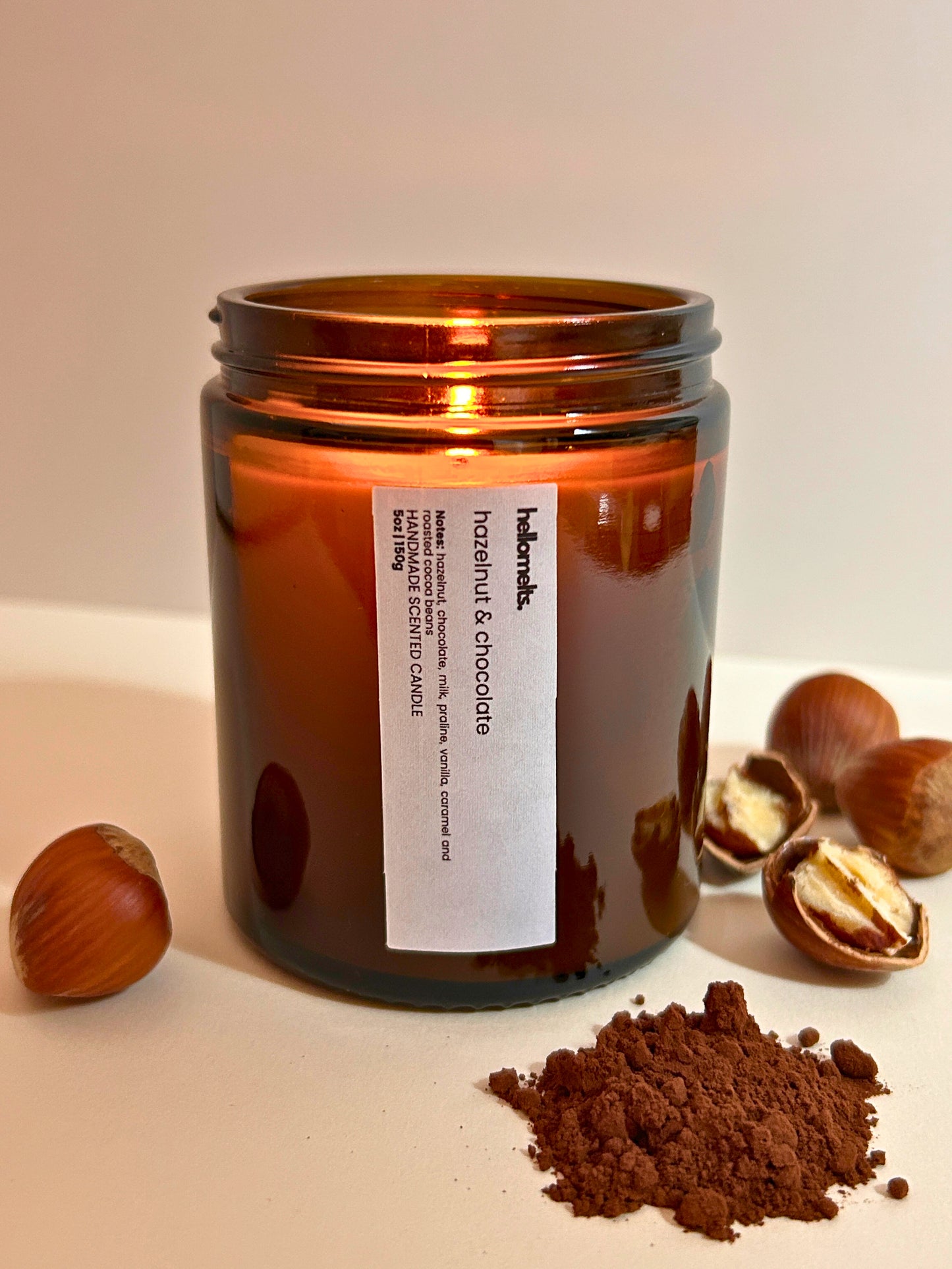 Hazelnut & Chocolate scented candle by Hellomelts, lit amber glass jar styled on a white background with whole hazelnuts and cocoa powder, showing a warm, rich chocolate and nut fragrance theme