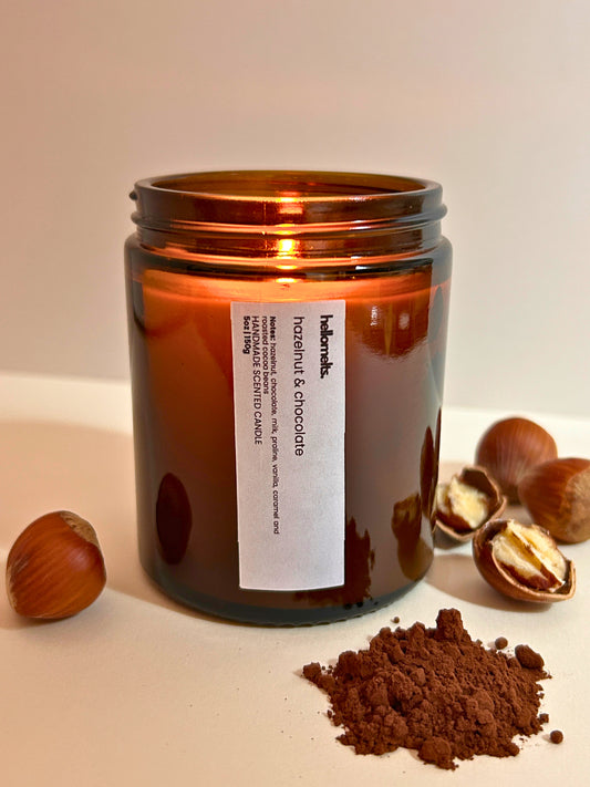 Hazelnut & Chocolate scented candle by Hellomelts, lit amber glass jar styled on a white background with whole hazelnuts and cocoa powder, showing a warm, rich chocolate and nut fragrance theme