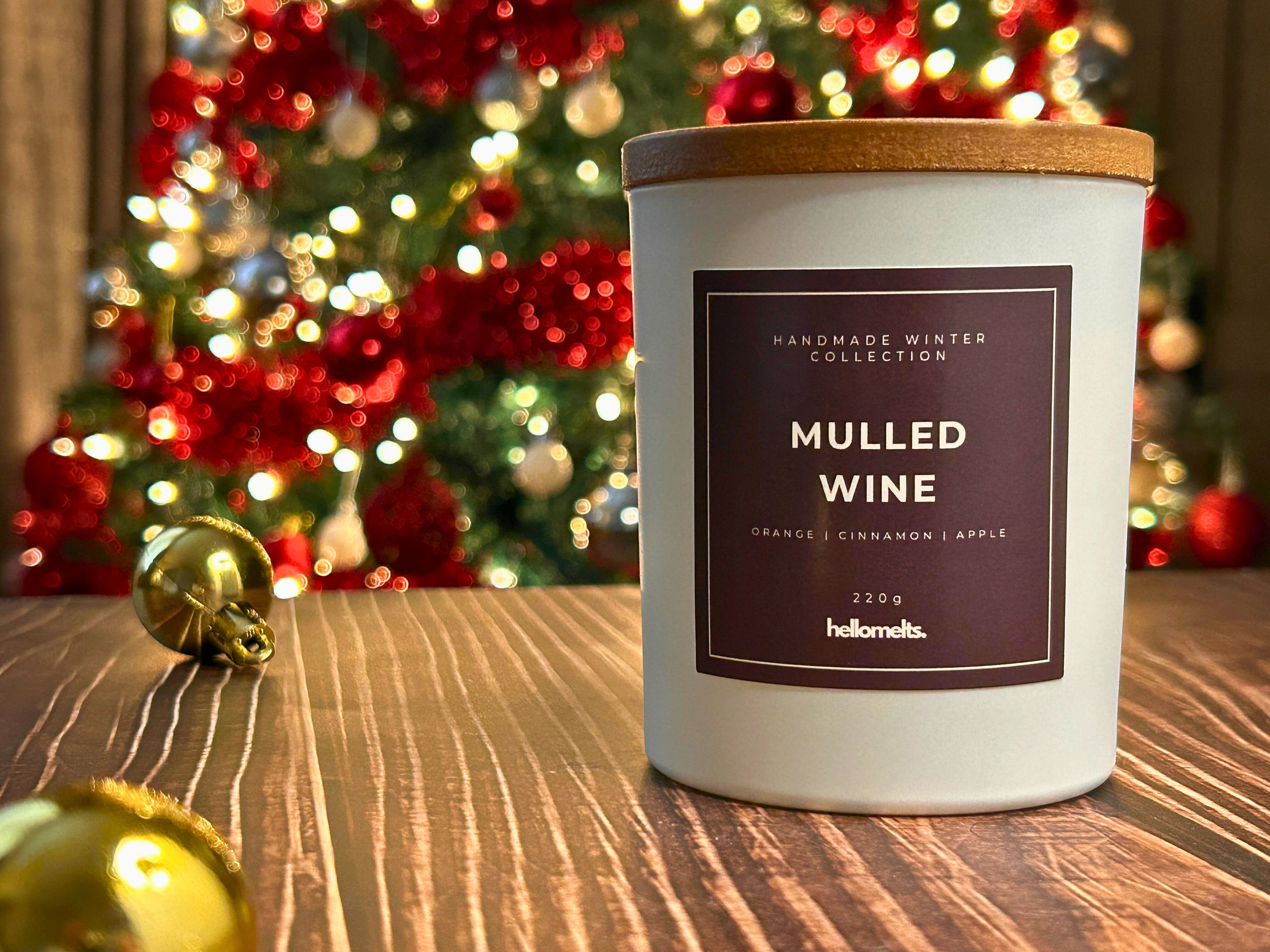 Mulled Wine winter candle by Hellomelts displayed on a wooden surface with a blurred Christmas tree and gold baubles in the background, creating a warm festive atmosphere