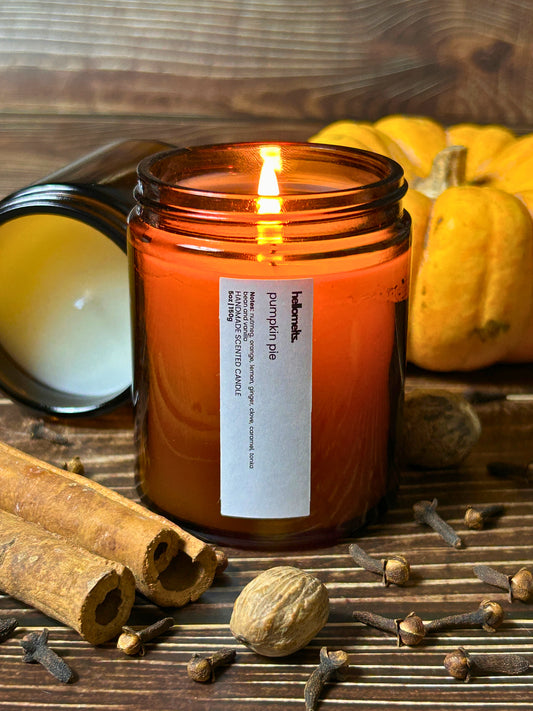 Pumpkin Pie scented candle by Hellomelts styled with cinnamon sticks, nutmeg, cloves and pumpkin on a rustic wooden surface, showcasing a warm spiced autumn fragrance