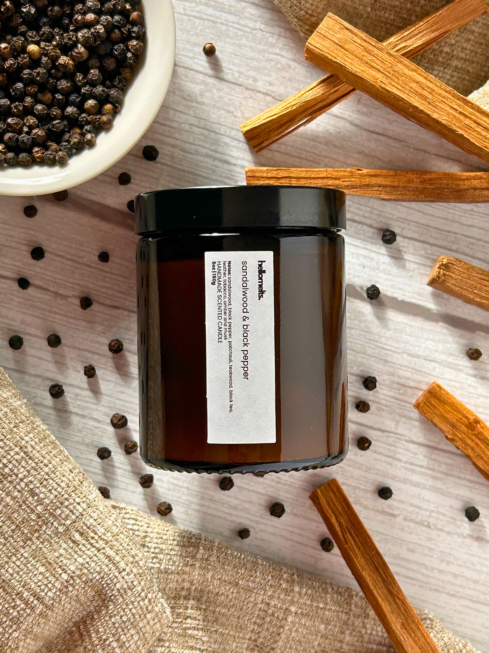Sandalwood & Black Pepper 150g scented candle by Hellomelts, styled in a warm lifestyle flatlay with peppercorns and sandalwood sticks, part of the Hellomelts Autumn Collection