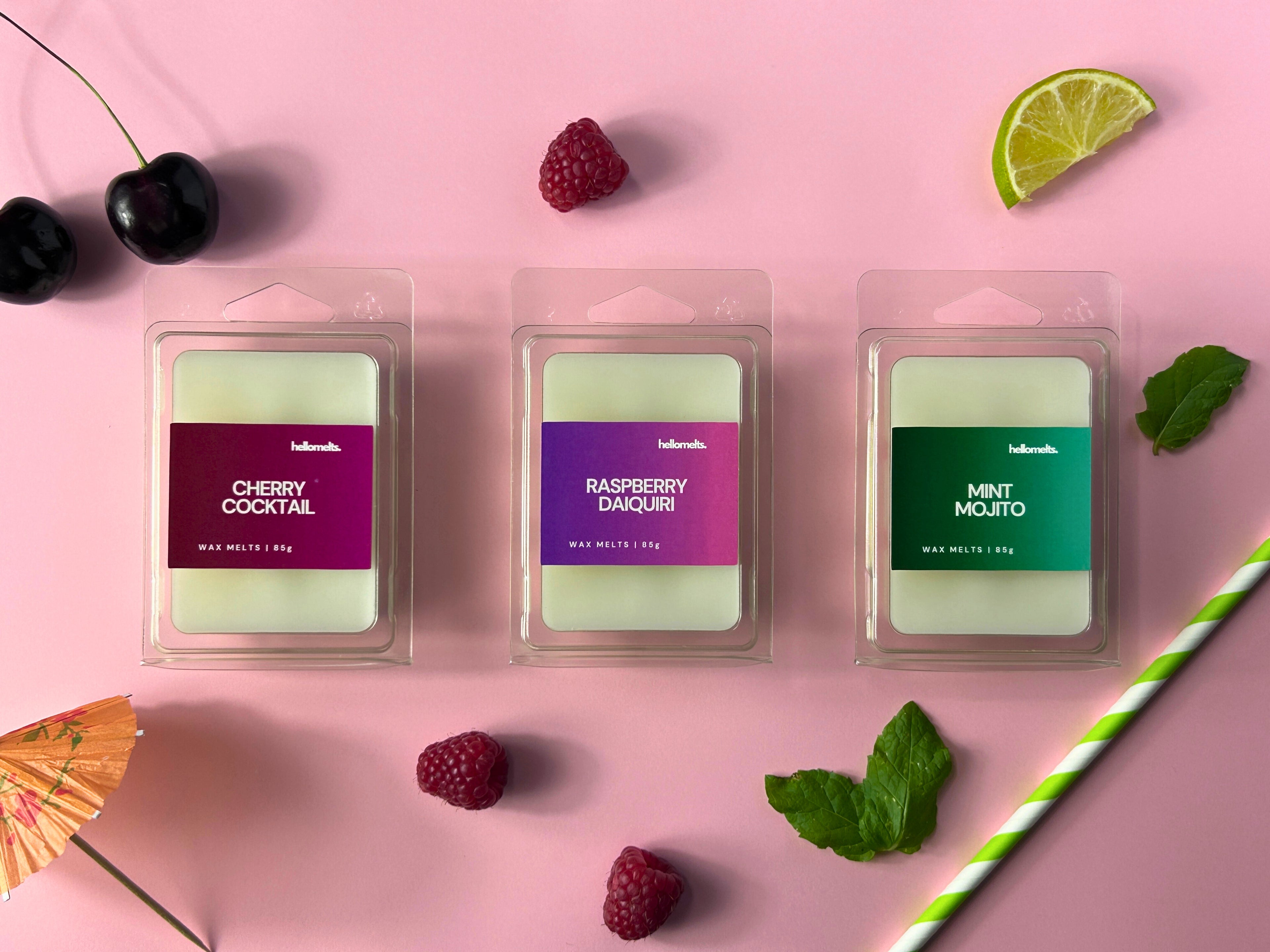 Flatlay of three Hellomelts 85g wax melt bars — Cherry Cocktail, Raspberry Daiquiri, and Mint Mojito — styled with fruit and cocktail props on a pink background, part of the Summer Collection